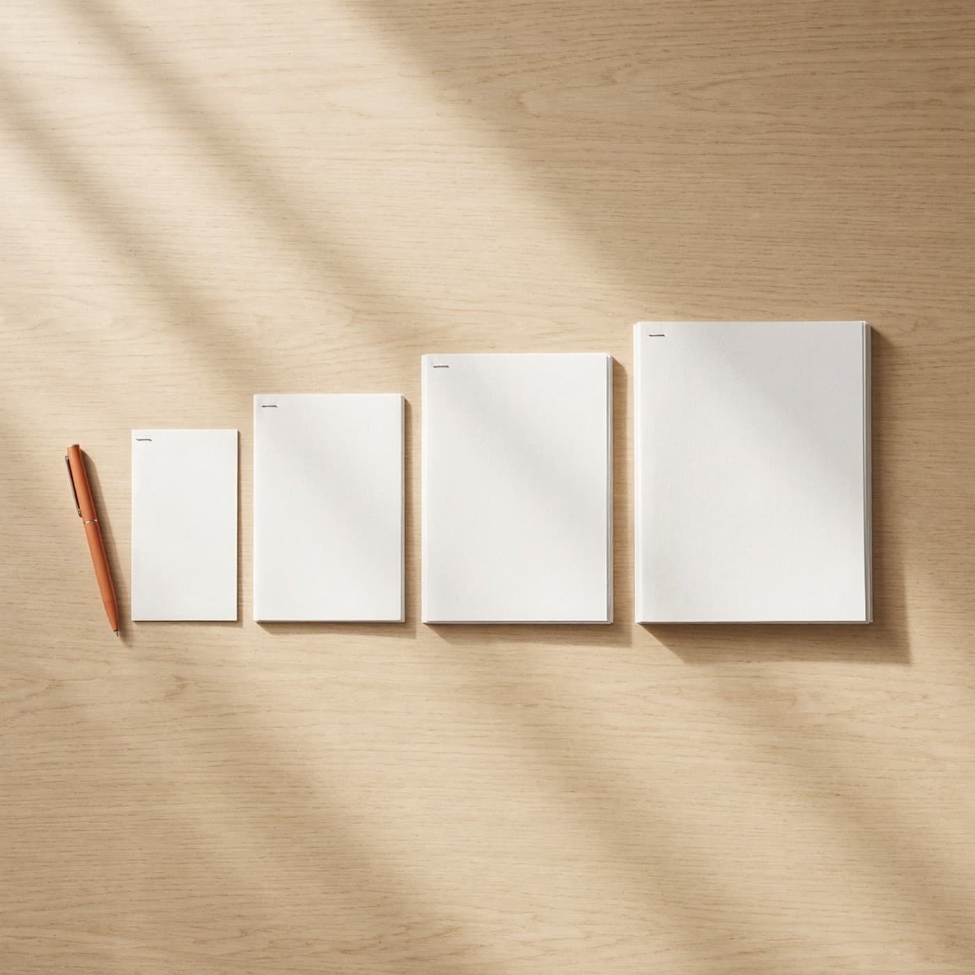 Four blank paper proposals laid out on an oak desk, each progressively thicker than the last, with a single orange pen beside the thinnest one
